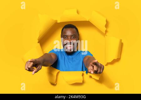 Joyeux afro-américain homme pointant les doigts vers l'appareil photo, vous choisissant, posant dans le trou déchiré de papier jaune arrière-plan Banque D'Images
