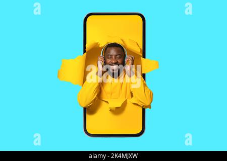 Homme afro-américain joyeux dans l'écran de smartphone, avec un casque sans fil et en écoutant de la musique avec les yeux fermés Banque D'Images