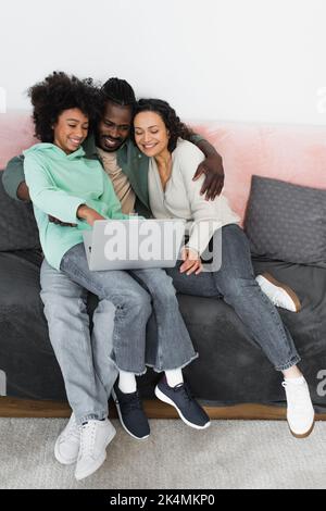 vue en grand angle d'une famille afro-américaine joyeuse regardant un film de comédie sur un ordinateur portable tout en étant assis sur un canapé, image de stock Banque D'Images