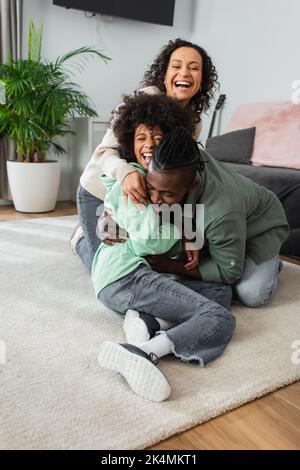 joyeux père afro-américain tickling la fille de la préadolescence près de la femme positive dans le salon, image de stock Banque D'Images