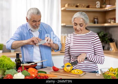 Des passe-temps de retraite. Homme senior avec smartphone capturant sa femme cuisine dans la cuisine Banque D'Images