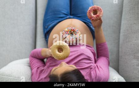 Femme enceinte mangeant des beignets sucrés sur le ventre. Fringales de desserts et de nourriture malsaine pendant la grossesse, drôle vue de dessus des gâteaux sur la bosse de bébé pour Banque D'Images