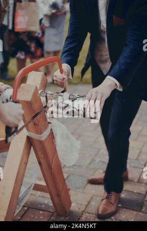 Un gros plan vertical du marié et de la mariée sciant un tronc d'arbre pendant la cérémonie de mariage Banque D'Images