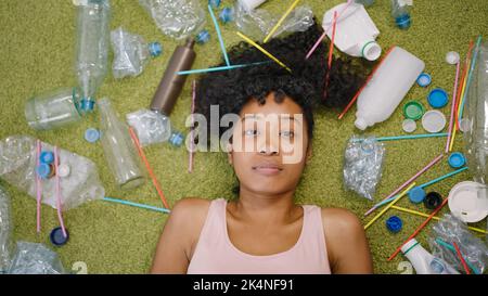 La femme afro-américaine repose sur un tapis vert moelleux près de déchets de plastique éparpillés. Une jeune femme de ménage au sol regarde dans la vue de dessus de la caméra de près Banque D'Images