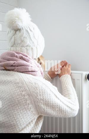 Une femme chauffe ses mains près de la batterie. Femme habillée chaleureusement froide dans la maison. Économie de chauffage. Factures de services publics coûteuses. Banque D'Images