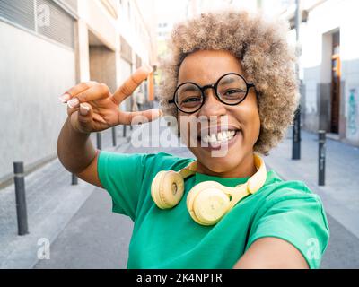 Perspective selfie d'une femme afro-américaine heureuse avec des cheveux afro portant des lunettes et des écouteurs dans la ville faisant le symbole de la paix Banque D'Images