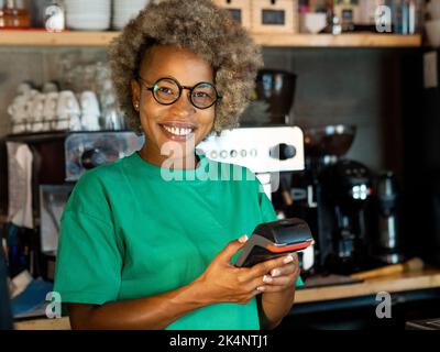 Serveuse afro-américaine souriante dans un pub avec une damaphone entre ses mains. Petites entreprises, paiements par carte Banque D'Images