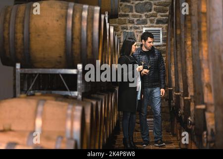 Couple dans la cave à vin, en appréciant boire du vin blanc tout en regardant les vieux fûts de bois pour le stockage et le vieillissement du vin Banque D'Images
