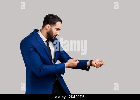 Vue latérale de l'homme barbu qui a l'air diligent et fort, debout dans l'attaque ou en tirant les mains gestuelle et regardant vers l'avant, en portant un costume de style officiel. Prise de vue en studio isolée sur fond gris. Banque D'Images