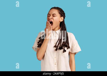 Jeune femme épuisée avec des dreadlocks noirs couvrant la bouche avec la main, garde les yeux fermés, sensation de fatigue ou de sommeil, portant une chemise blanche. Studio d'intérieur isolé sur fond bleu. Banque D'Images