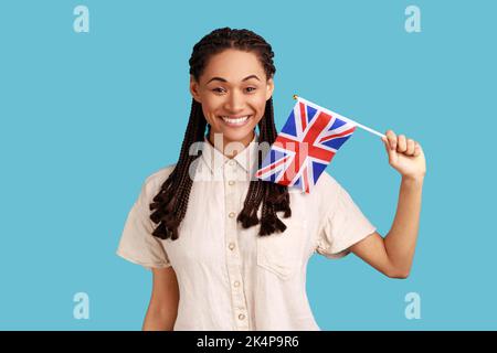 Portrait d'une femme souriante et satisfaite avec des dreadlocks noirs qui enferme le drapeau britannique, célébrant les vacances, regardant l'appareil photo, portant une chemise blanche. Studio d'intérieur isolé sur fond bleu. Banque D'Images