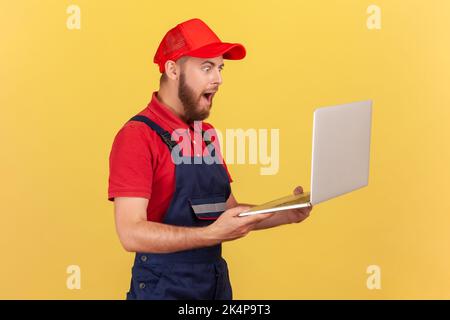 Portrait de côté d'un homme de main stupéfiant, debout en mains et travaillant en ligne, en train de passer un énorme ordre, en portant un uniforme et une casquette rouge. Studio d'intérieur isolé sur fond jaune. Banque D'Images