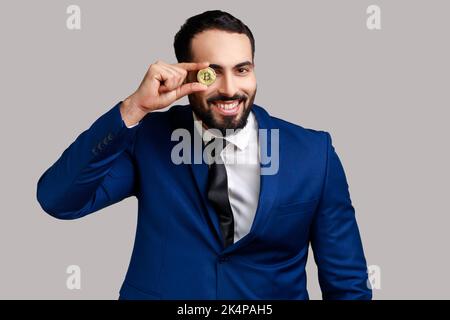 Portrait d'homme d'affaires barbu couvrant l'œil avec un bitcoin doré, regardant l'appareil photo avec un sourire éclatant, portant un costume de style officiel. Prise de vue en studio isolée sur fond gris. Banque D'Images