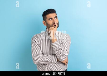 Portrait d'un homme d'affaires barbu et attentionné tenant son menton et méditant sur l'idée, confus pas sûr de la solution, portant une chemise à rayures. Studio d'intérieur isolé sur fond bleu. Banque D'Images