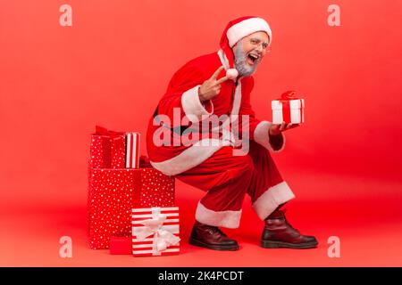 Homme âgé positif et excité avec barbe grise en costume du père noël assis sur une grande boîte avec cadeau de Noël, tenant le cadeau, montrant le signe v à l'appareil photo. Studio d'intérieur isolé sur fond rouge. Banque D'Images