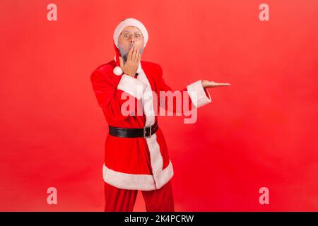 Homme âgé choqué avec une barbe grise portant le costume du père noël présentant un espace de copie sur sa paume, couvrant la bouche avec la main, expression étonnante. Studio d'intérieur isolé sur fond rouge. Banque D'Images