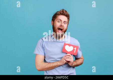 Portrait de l'homme barbu excité tenant le cœur des médias sociaux comme bouton devant sa poitrine, compteur d'emoji, notification de suiveur. Studio d'intérieur isolé sur fond bleu. Banque D'Images