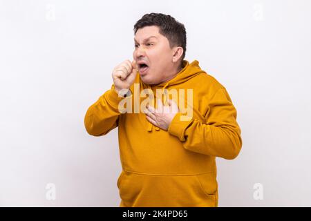 Portrait d'un homme d'âge moyen malsain toussant, attrape froid, ayant une température élevée, ayant un symptôme de grippe, portant un pull à capuche de style urbain. Studio d'intérieur isolé sur fond blanc. Banque D'Images