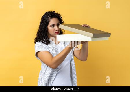 Portrait d'une femme triste déçue avec des cheveux ondulés foncés debout avec boîte cadeau dans les mains, regardant à l'intérieur et n'aime pas son cadeau pour l'anniversaire. Studio d'intérieur isolé sur fond jaune. Banque D'Images