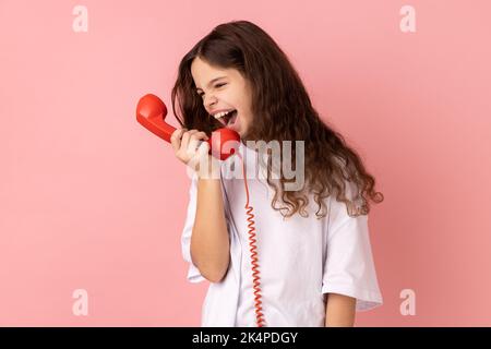 Portrait de la colère agressive belle petite fille portant un T-shirt blanc criant dans le téléphone rétro combiné, irrité par l'appel. Studio d'intérieur isolé sur fond rose. Banque D'Images