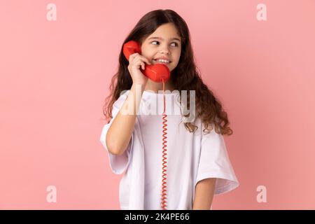 Portrait d'une petite fille satisfaite portant un T-shirt blanc parlant téléphone fixe tenant dans le combiné, regardant loin avec un sourire crasseux. Studio d'intérieur isolé sur fond rose. Banque D'Images