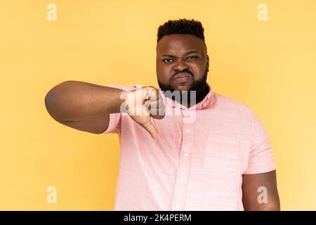 Portrait de triste bouleversé barbu homme portant une chemise rose montrant le pouce vers le bas, exprimant de mauvaises émotions négatives, n'aime pas, frognant visage. Studio d'intérieur isolé sur fond jaune. Banque D'Images