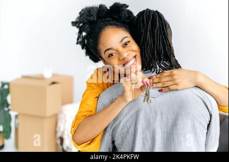 Investissement, concept de propriété. Positive excitée femme afro-américaine embrassant son mari en se tenant dans le salon, elle montre les clés de leur nouvelle propriété, regarde la caméra, souriant heureux Banque D'Images