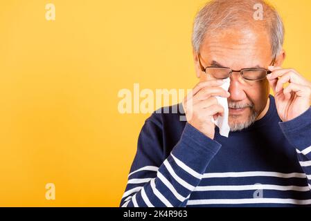 Asiatique homme aîné pleure lever des lunettes avec tissu essuyant les yeux rouges studio tourné isolé sur fond jaune, Portrait vieux homme triste essuyant son Banque D'Images