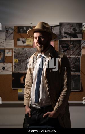 Portrait d'un détective privé pensif dans un manteau de tranchée et un chapeau près du tableau des preuves dans le bureau la nuit dans le bureau. Banque D'Images