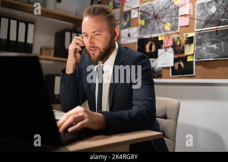 Beau policier assis à côté de l'ordinateur, parlant sur un téléphone cellulaire, tableau de preuve sur fond Banque D'Images
