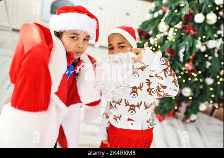 Enfants - frère et soeur dans un grand costume de santa avec une fausse barbe jouer et grimace sur le fond de l'arbre de Noël. Banque D'Images