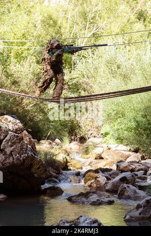 Un militaire ou un joueur d'aérosoft dans un costume de camouflage se faufilant sur le pont de corde et vise d'un fusil de sniper sur le côté ou à la cible. Banque D'Images