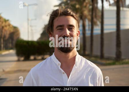 Photo gaie d'un beau jeune homme caucasien avec de longs cheveux blonds regardant la caméra, à l'extérieur. Banque D'Images