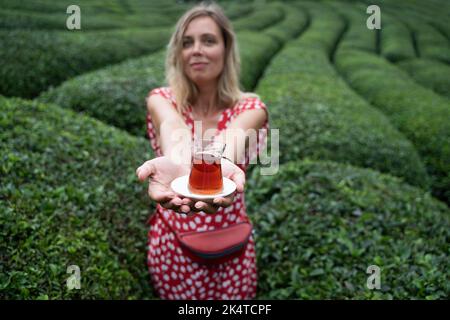 Vue de face de la jeune femme en robe rouge à pois offrant un verre traditionnel de thé noir turc frais avec des rangées de plantations de thé Banque D'Images