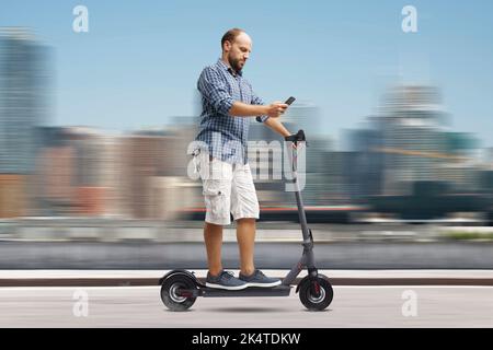 Homme imprudent utilisant un smartphone tout en conduisant un scooter électrique, ville en arrière-plan, concept de mobilité durable Banque D'Images