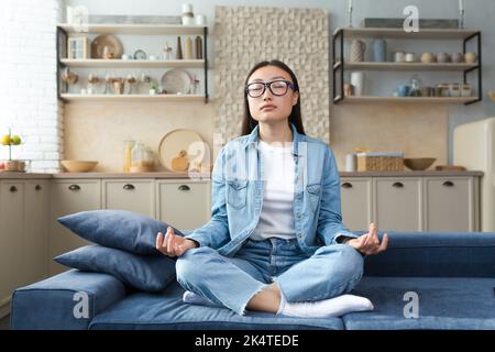 Belle femme asiatique se détendant à la maison assis sur un canapé et méditant dans la pose de lotus, faisant des exercices de respiration dans le salon. Banque D'Images