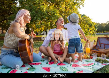 Famille de quatre avec maman jouant de la guitare assis pique-nique dans le parc. Banque D'Images