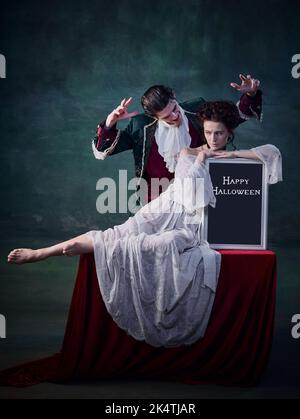 Portrait de la jeune belle femme en robe blanche assise sur la table avec le comprimé d'Halloween et l'homme, vampire la craquant Banque D'Images