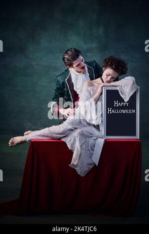 Portrait de la jeune belle femme en robe blanche assise sur une table avec un comprimé d'Halloween et un homme de caractère de vampire la mordant Banque D'Images