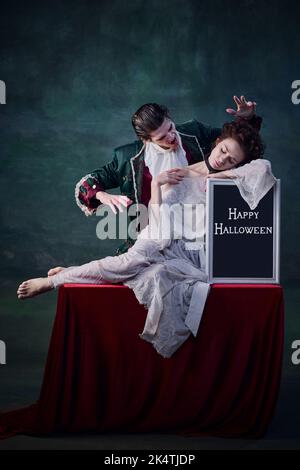 Portrait de la jeune belle femme en robe blanche assise sur une table avec un comprimé d'Halloween et un homme de caractère de vampire la mordant Banque D'Images