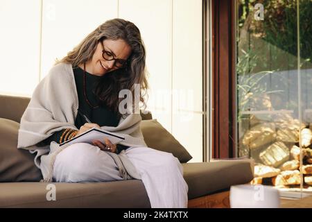 Journalisation pendant la retraite. Femme mûre souriant en écrivant dans son journal à la maison. Une femme âgée gaie qui écrit ses pensées pendant Banque D'Images