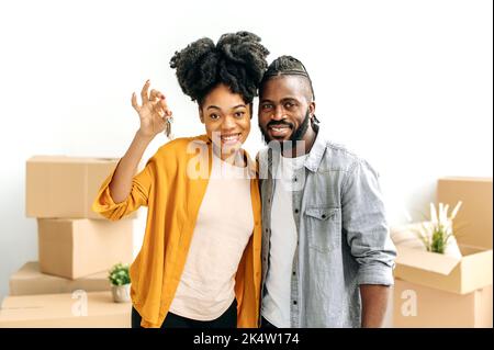 Heureux marié african american couple, newlyweds, a acheté leur première maison, se tenir dans la salle de séjour contre le fond de boîtes en carton avec des choses, regarder l'appareil photo, sourire, la fille montre les clés Banque D'Images