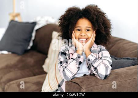 Portrait de petite fille afro-américaine mignonne et gaie d'âge préscolaire avec des cheveux bouclés, dans des vêtements décontractés, assis sur le canapé dans le salon, pose pour l'appareil photo, souriant heureux Banque D'Images