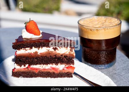 Une tranche de gâteau au chocolat aux fraises de Devils frais servi sur une table de café extérieure. le gâteau à la crème fraîche est servi avec un verre de café glacé Banque D'Images