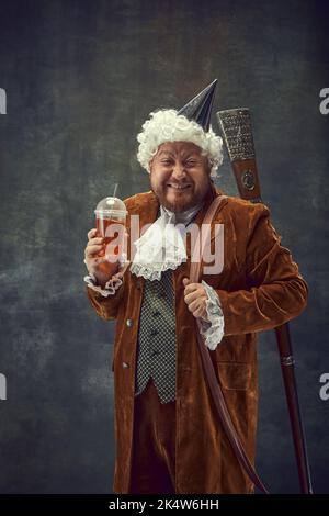 Drôle de jeune homme en costume brun vintage et perruque blanche comme chasseur royal médiéval avec vieille arme de rire isolé sur fond sombre. Banque D'Images