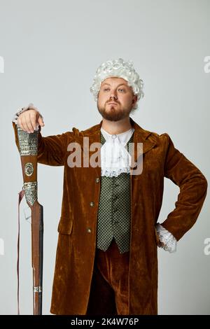 Honneur. Photo de studio d'un jeune homme à l'image d'une personne médiévale en costume de chasse brun vintage et perruque blanche avec ancien fusil de chasse isolé sur gris Banque D'Images