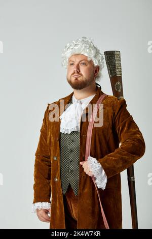 Photo de studio d'un jeune homme à l'image d'une personne médiévale en costume de chasse brun vintage et perruque blanche avec ancien fusil de chasse isolé sur fond gris. Banque D'Images