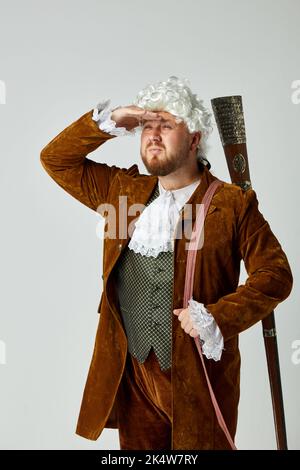 Recherche de proies. Jeune homme à l'image d'une personne médiévale en costume de chasse brun vintage et perruque blanche avec ancien fusil de chasse isolé sur gris Banque D'Images