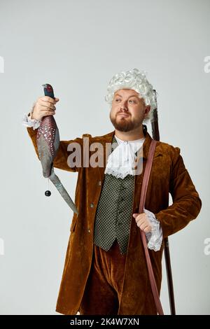 Photo de studio d'un jeune homme à l'image d'une personne médiévale en costume de chasse brun vintage et perruque blanche avec ancien fusil de chasse isolé sur fond gris. Banque D'Images