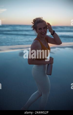 femme de fitness souriante jogger dans des vêtements de fitness avec une bouteille d'eau marchant à la plage au coucher du soleil. Banque D'Images